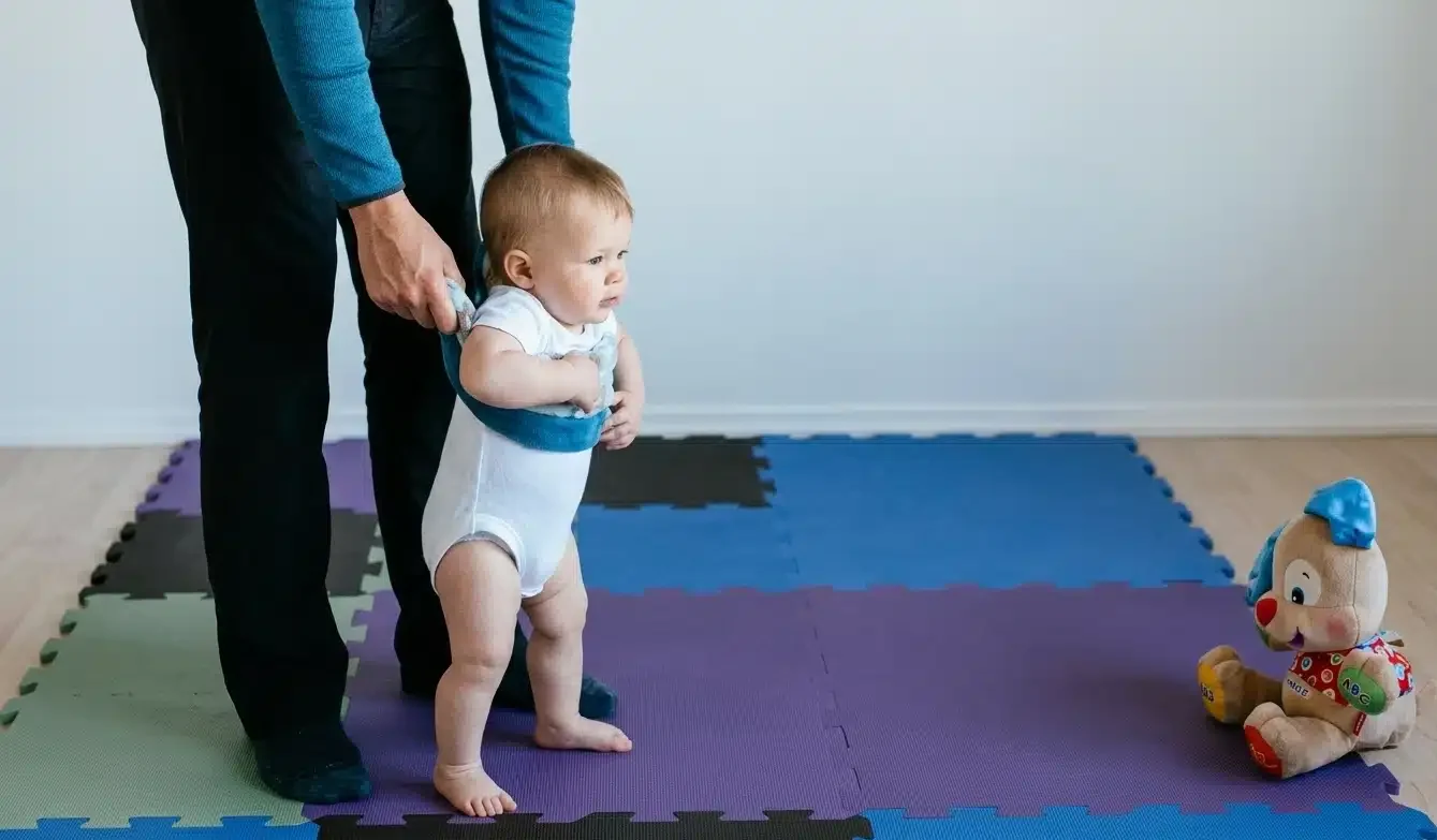 A professional nanny gently guides a clothed baby, holding him by the armpits, through floor movements on soft rugs. This is efficient nanny walking methods