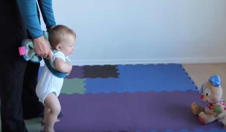 A professional nanny gently guides a clothed baby, holding him by the armpits, through floor movements on soft rugs. This is efficient nanny walking methods.