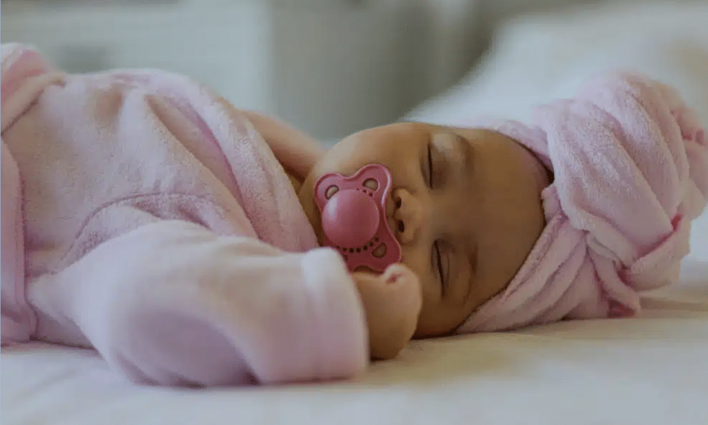A calm newborn baby sleeping in a pink outfit with a pacifier. Early signs of pacifier-related damage.